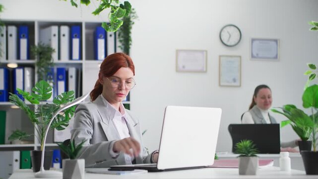 working atmosphere, young woman in glasses busy with work writing reports background of female colleague slacking off in office on a working day