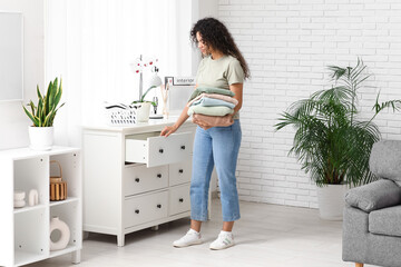 Beautiful young African-American woman putting clean clothes into commode at home