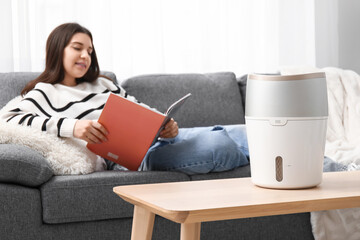 Young woman reading magazine on sofa near modern humidifier in living room