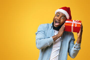 Portrait of crazy black guy in Santa hat holding Christmas gift box, excited about what's inside, yellow studio background with free space