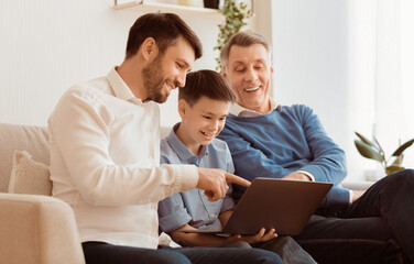 Boy Using Laptop Sitting Between Father And Grandpa Doing Homework Online On Sofa At Home. Selective Focus