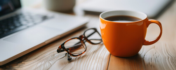 A bright orange coffee mug sits next to a laptop and glasses on a wooden desk, creating a cozy workspace.