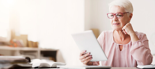 Smiling beautiful senior woman with glasses using digital tablet at home, empty space