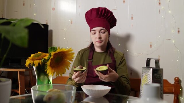 Lady in red chef hat and apron is dividing ripe avocado in to halves and extracting seed filmed in slow motion. Caucasian woman is preparing healthy green product sitting at the table in cozy kitchen.