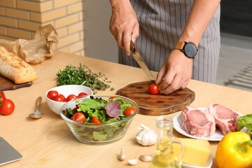 Young man cutting tomato at table in kitchen, closeup