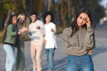 Sad muslim girl staying aside from laughing international group of teenagers, park background