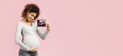 Happy afro pregnant woman with her baby sonography, enjoying first photo of her unborn child, pink...