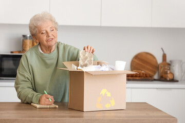 Senior woman with notebook and box of garbage in kitchen. Waste sorting concept