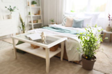 Blurred view of light bedroom with tables, shelf unit and plants