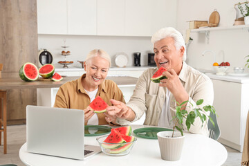 Mature couple with slices of fresh watermelon video chatting at table in kitchen