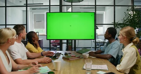 A group of young people are looking at a green screen while sitting at a table in an office meeting room. Copy space, mockup - Powered by Adobe