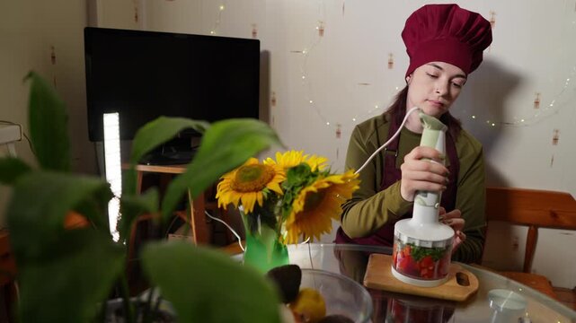 Woman is using hand blender to cook guacamole sauce sitting at the kitchen table. Slow motion footage of lady in chef hat preparing delicious vegetarian meal using electronic mixer. Nourishment theme.