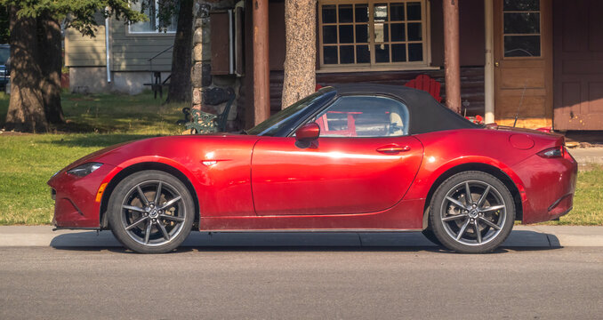 Waterton Park, AB, Canada. Aug 20, 2024. A vibrant red Mazda MX-5 Miata convertible gleams in the sunlight, parked alongside a charming wooden house with a backdrop of lush green trees.