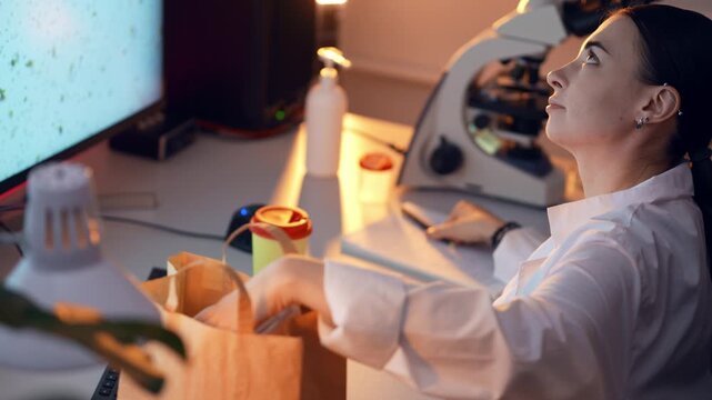 Woman is taking out lunch boxes from paper bag sitting at the desk in front of computer in laboratory. Female Caucasian scientist is going to eat food in slowmo. Having break for meal at the job.