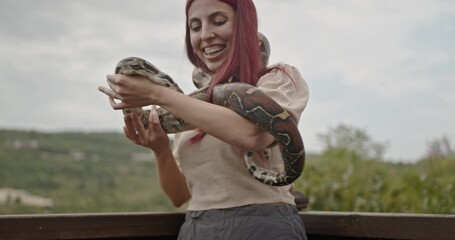 A happy young adult female plays with her pet snake outdoors. She has vibrant red hair and a big smile.