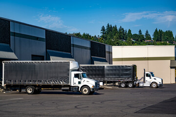 Day cab white big rig semi trucks with Conestoga trailers loading cargo standing in warehouse dock...