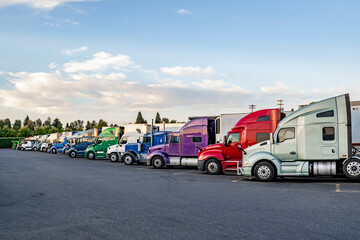 Long row of the big rig semi trucks with semi trailers standing on the truck stop parking lot at twilight time