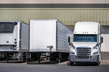 White long haul big rig semi truck with semi  trailer and another refrigerator semi trailers loading cargo standing in warehouse dock gates