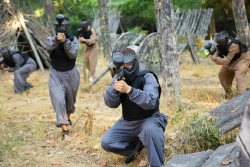 Fototapeta premium Group of men and women in protective clothes and helmets shooting with paintball markers. Teams fighting on battlefield outdoors.