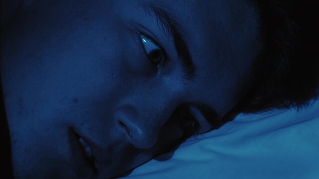 Nightmare fear. Sleep disorder. Disturbed scared frightened guy woke up opened eyes in bed late at night in dark blue light.