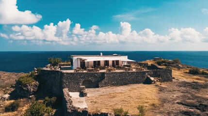 Aerial shot of a white-brick cafe, stone walls surrounding it, set on a remote island under blue skies and white clouds with the sea in the distance.