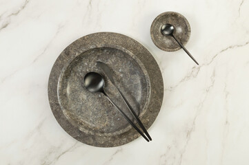 Top view of brown marble tray, plate, bowl and black cutlery on white marble background. Food presentation, dishes, stone tray. Copy space, flat lay.