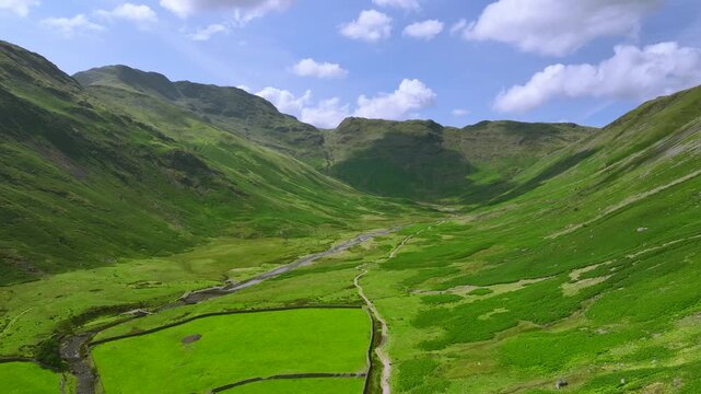 Flying above wide green valley surrounded by mountains with stream and path. Mickleden, The Langdales, Lake District, UK