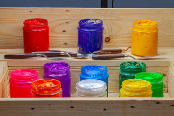  A colorful arrangement of open acrylic paint jars showcasing a spectrum of vibrant hues, from warm reds and oranges to cool blues and greens. The wooden box and palette knives in the background.