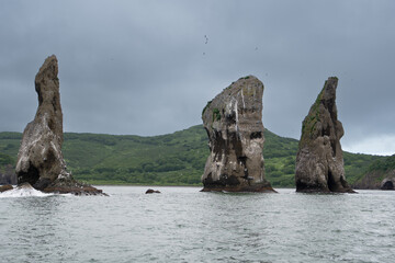From the sea side there are Three brothers rocks in the Avacha Bay. In the background there are green mountains and hills.