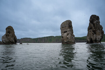 From the sea side there are Three brothers rocks in the Avacha Bay. In the background there are green mountains and hills.