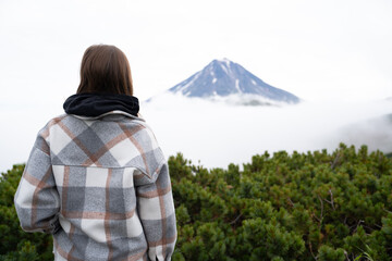 The large Vilyuchinsky volcano has an edge visible in clouds and fog. In the foreground is a cedar elfin and there is a girl in a brown poncho looking at the 