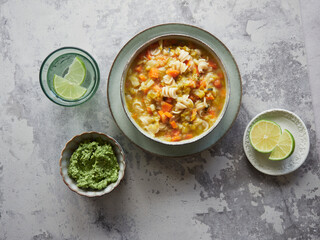 top view of plate of delicious vegetable and pasta soup called minestrone served on gray countertop with green pesto sauce, glass of water and lime slices.