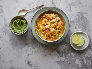 delicious vegetable and pasta soup called minestrone served on a grey countertop with green pesto sauce, antique spoon and lime slices, top view