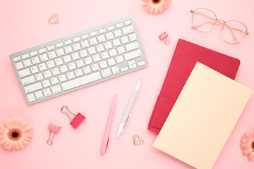 Elegant feminine workspace with keyboard, paper notebooks, clips, pens, flowers, eyeglasses on pastel pink background. Distance job, work at home concept.
