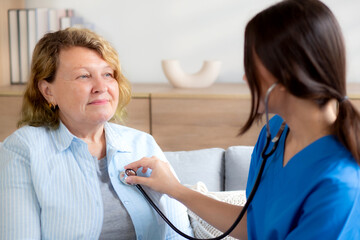 Caregiver young nurse woman examining elderly with stethoscope for checkup sitting on sofa in living room at home, nurse checkup patient about cardiac, patient and insurance, medical and health.