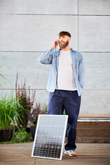 Bearded man talks on smartphone connected to photovoltaic solar panel. Integration of sustainable...