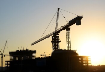 Obraz premium A large construction crane silhouetted against a sunset sky, with the outline of a building under construction in the background