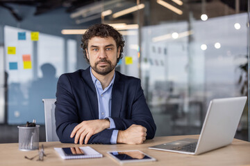 Confident businessman in a modern office environment, seated at a desk with laptop and tablet, exuding professionalism and determination.