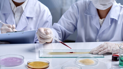 A yellow hair sample on a glass tray is being dropped by the scientist drop by drop of a red substance. The remaining scientist is carefully taking notes. Scene of hair research in the laboratory