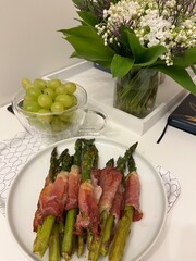 green asparagus wrapped in cured ham on a white plate, a bunch of grapes in a cup and a bouquet in a vase