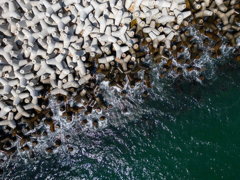 Aerial view of concrete tetrapod wave breakers along a rugged shoreline, with waves crashing against them, creating a dynamic and textured seascape. Ideal for coastal, industrial, and nature themes