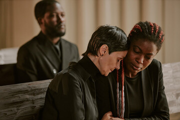 African American daughter supporting her senior mother during funeral while her brother listening...