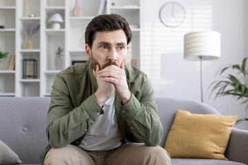 Thoughtful man in casual clothes sits on couch in modern living room, lost in contemplation. Calm atmosphere signifies introspection, choice-making. Emotion conveyed is pensive, thoughtful reflection