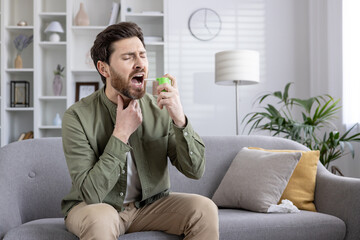 Man sitting on gray couch using throat spray to relieve soreness. Appears to be experiencing...