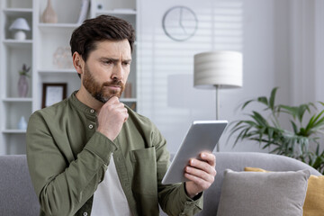 Thoughtful man holding tablet while sitting on sofa in cozy living room. Casual attire and pensive expression suggest contemplation or online browsing.