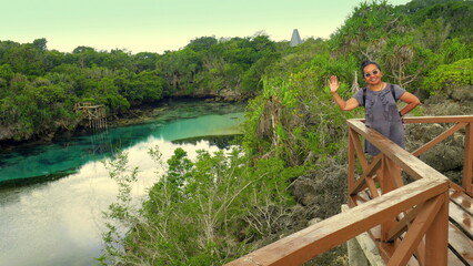 Fototapeta premium lächelnde Asiatin winkt fröhlich vom Aussichtssteg der blauen Lagune im Westen der Insel Sumba in tropischer Landschaft