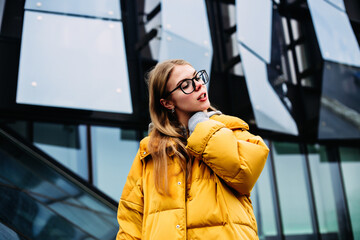 A blonde girl in a yellow jacket walks around the city. Girl with glasses