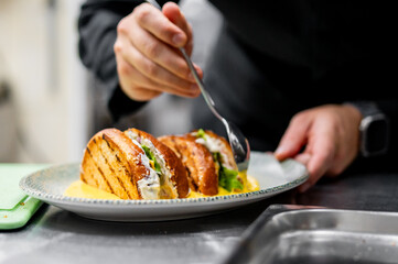 A close-up of hands garnishing a sandwich with sauce using a spoon. The sandwich is on a plate on a metal surface, suggesting a kitchen setting. Ideal for culinary content or food presentation themes.