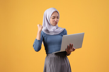 asian indonesian muslim woman holding laptop computer with angry face expression on isolated yellow background