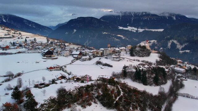 A scenic aerial view of a charming snow-covered village nestled in the mountains, surrounded by dramatic peaks under a cloudy winter sky.